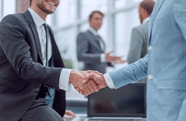 Two business professionals shaking hands in a modern office setting with colleagues in the background.