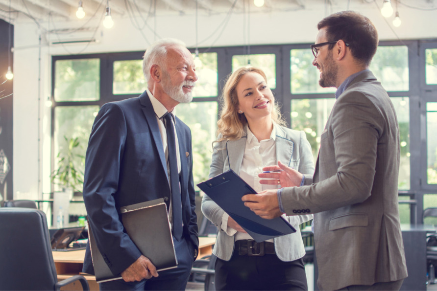 Three business professionals having a discussion in a modern office environment, holding documents and wearing suits.
