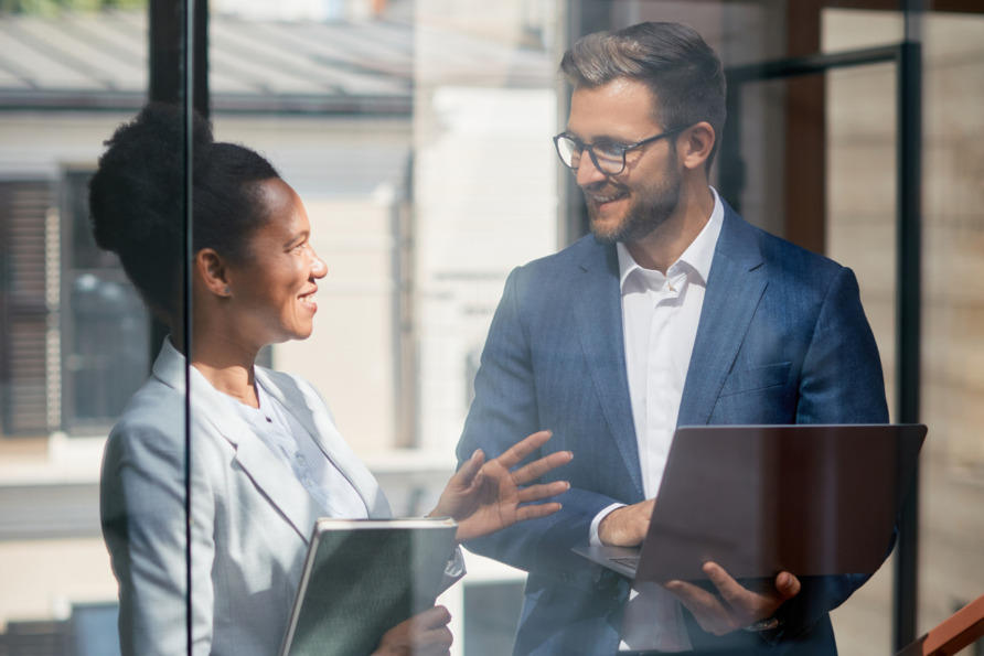 Two professionals in business attire having a discussion, one holding a notebook and the other with a laptop, by a window.