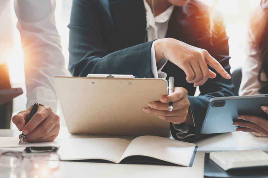 Professionals discussing documents at a meeting table, one pointing at a tablet, papers and pen visible.