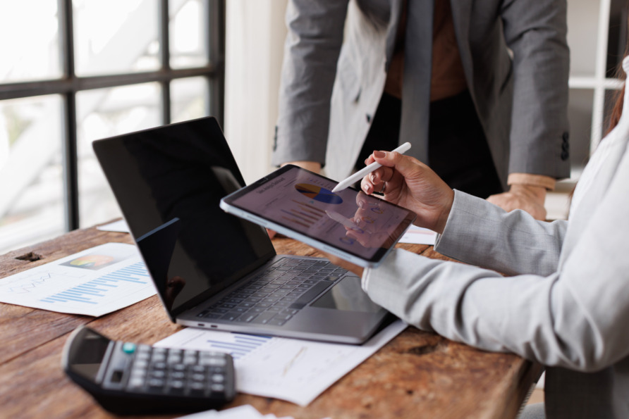 Person in suit holding a tablet with charts, working at a desk with a laptop, documents, and calculator nearby.