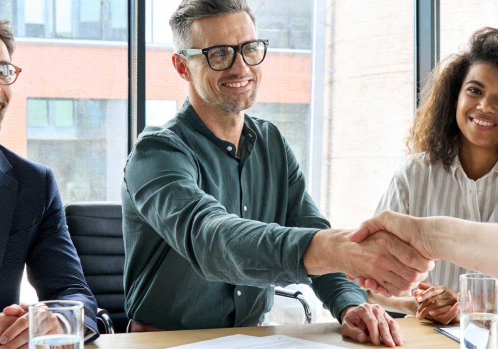 Four people in a business meeting, two shaking hands across a table, smiling colleagues watching in a modern office setting.