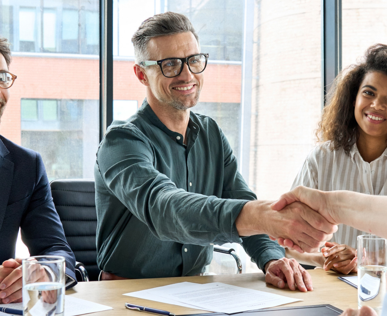 Four people in a business meeting, two shaking hands across a table, smiling colleagues watching in a modern office setting.