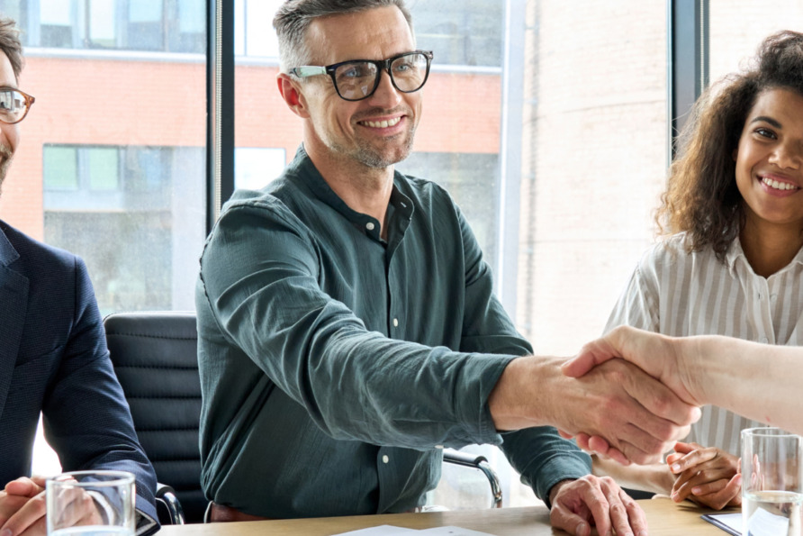 Four people in a business meeting, two shaking hands across a table, smiling colleagues watching in a modern office setting.