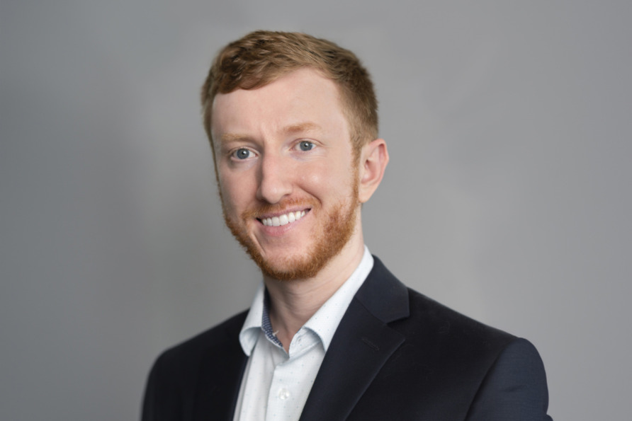 Man in a black suit with a light blue shirt, smiling against a gray background.